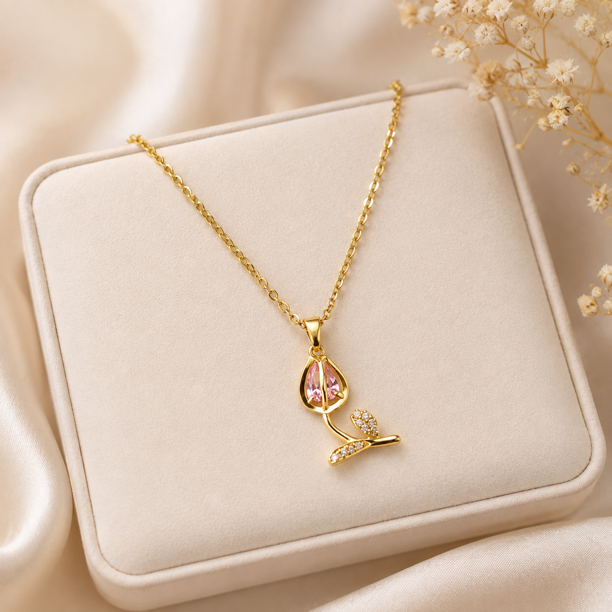 A gold plated anti tarnish tulip necklace with a teardrop-shaped pink stone and a small leaf detail, displayed on a cream-colored jewelry box