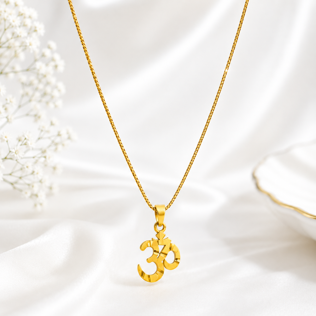 Celeste gold plated Om Necklace, displayed on a white satin fabric background with small white flowers on the left.