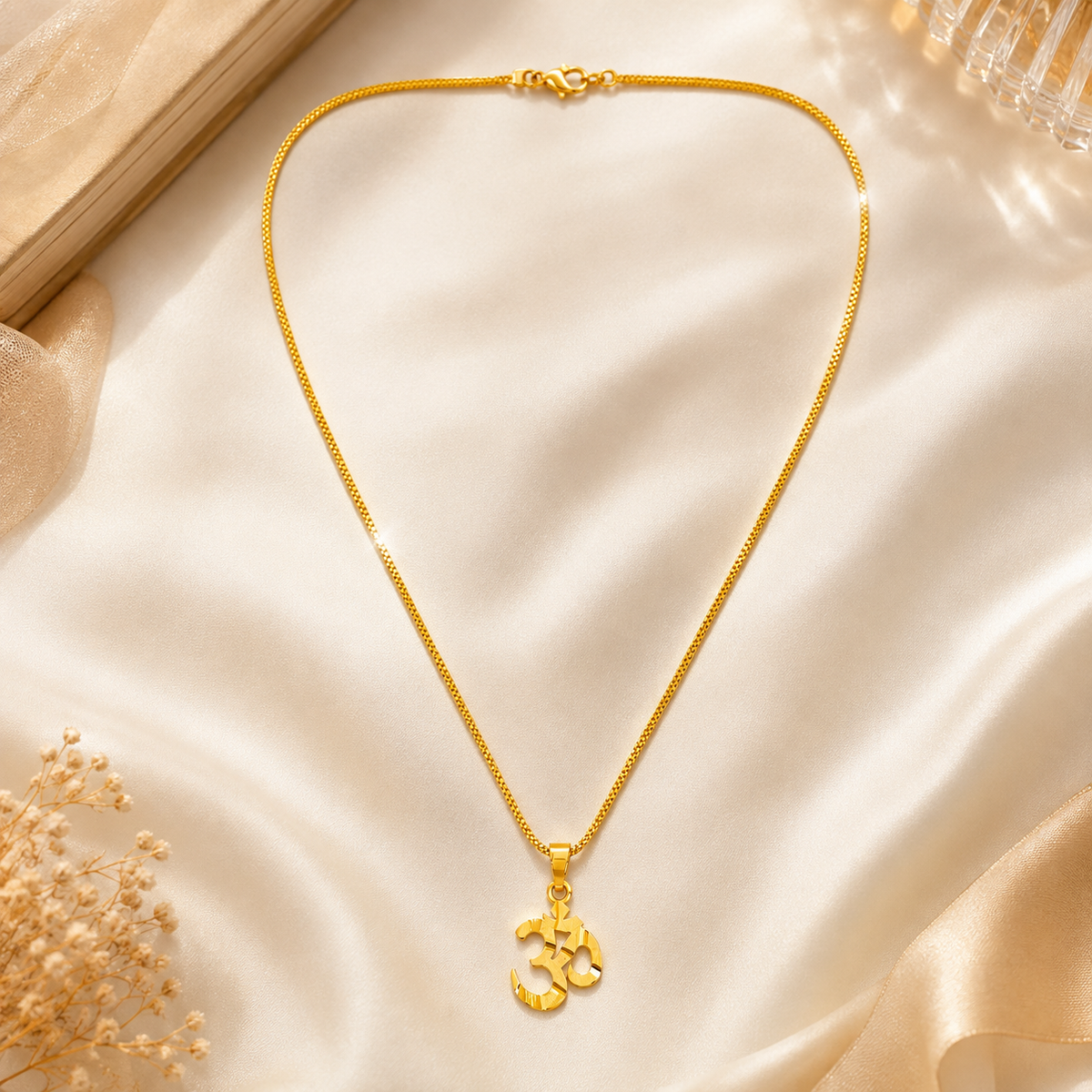Celeste gold plated Om Necklace, displayed on a draped cream satin fabric with decorative dried flowers